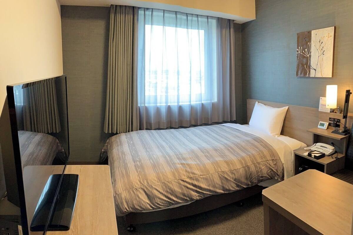 A hotel room with a bed, a desk, a chair, a nightstand with a lamp, and a painting on the wall.