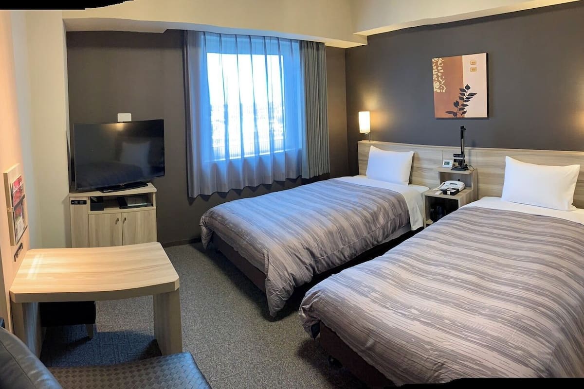 A hotel room with two beds, a desk, a television, and a window with curtains.
