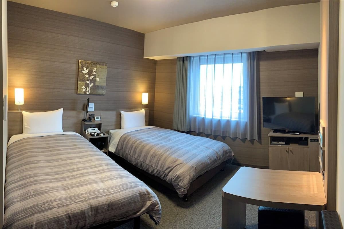 A hotel room with two beds, a desk, a television, and a window with curtains.