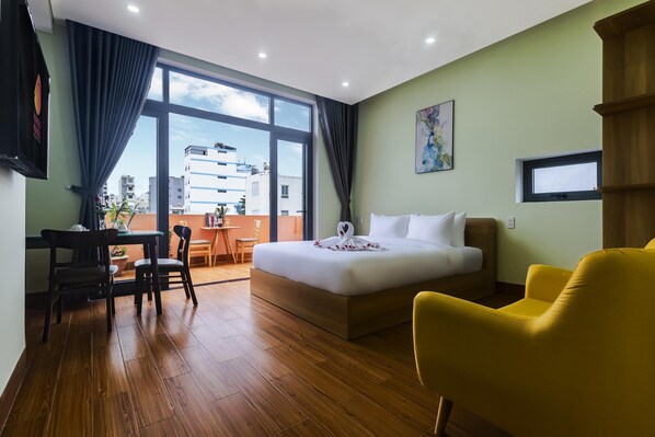 Une chambre d’hôtel avec un lit, un bureau, des chaises, un fauteuil jaune, une fenêtre donnant sur des immeubles et un tableau accroché au mur.