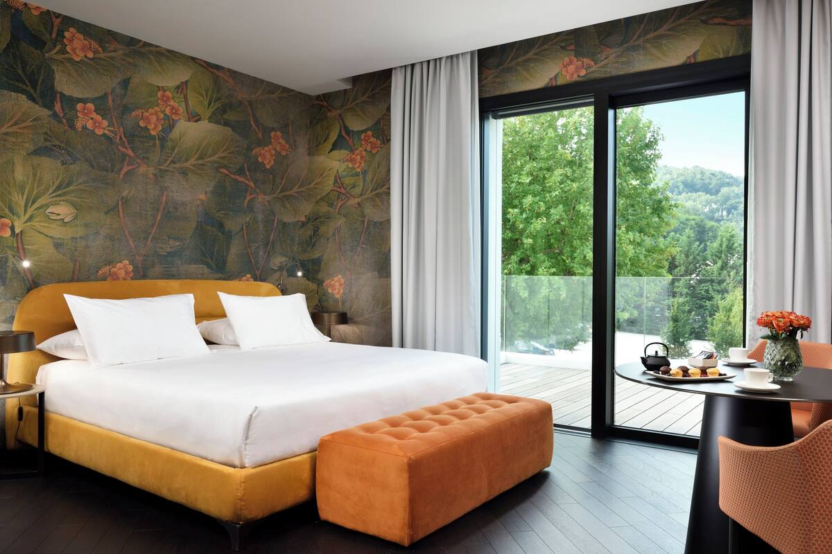A modern bedroom with a large bed, a wooden bench, a dining table with a vase, and a balcony with a view of greenery.