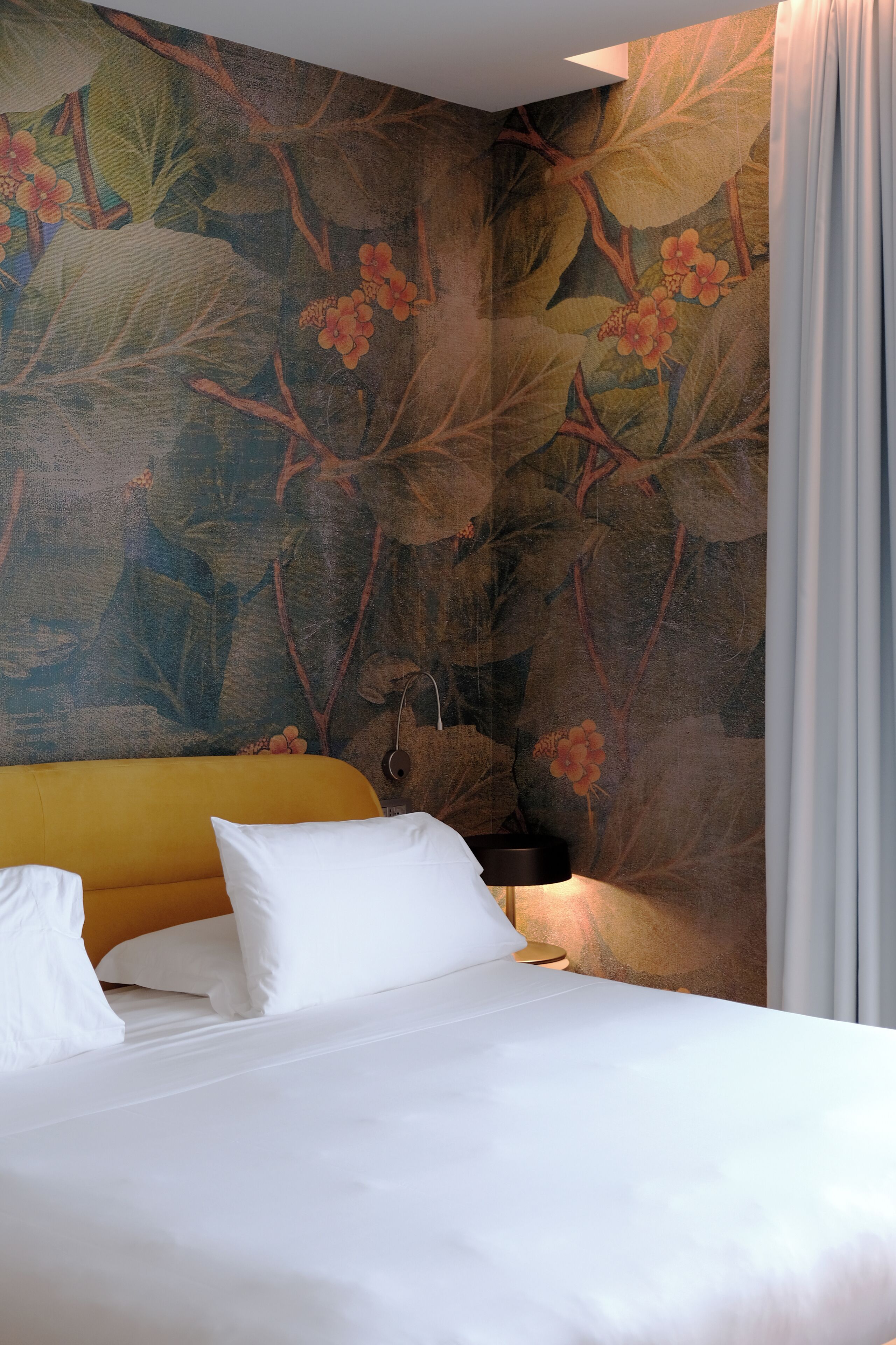 Ein Hotelzimmer mit Bett, gelbem Kopfteil, einer großen Wandmalerei mit floralem Muster und einem Nachttisch mit Lampe.