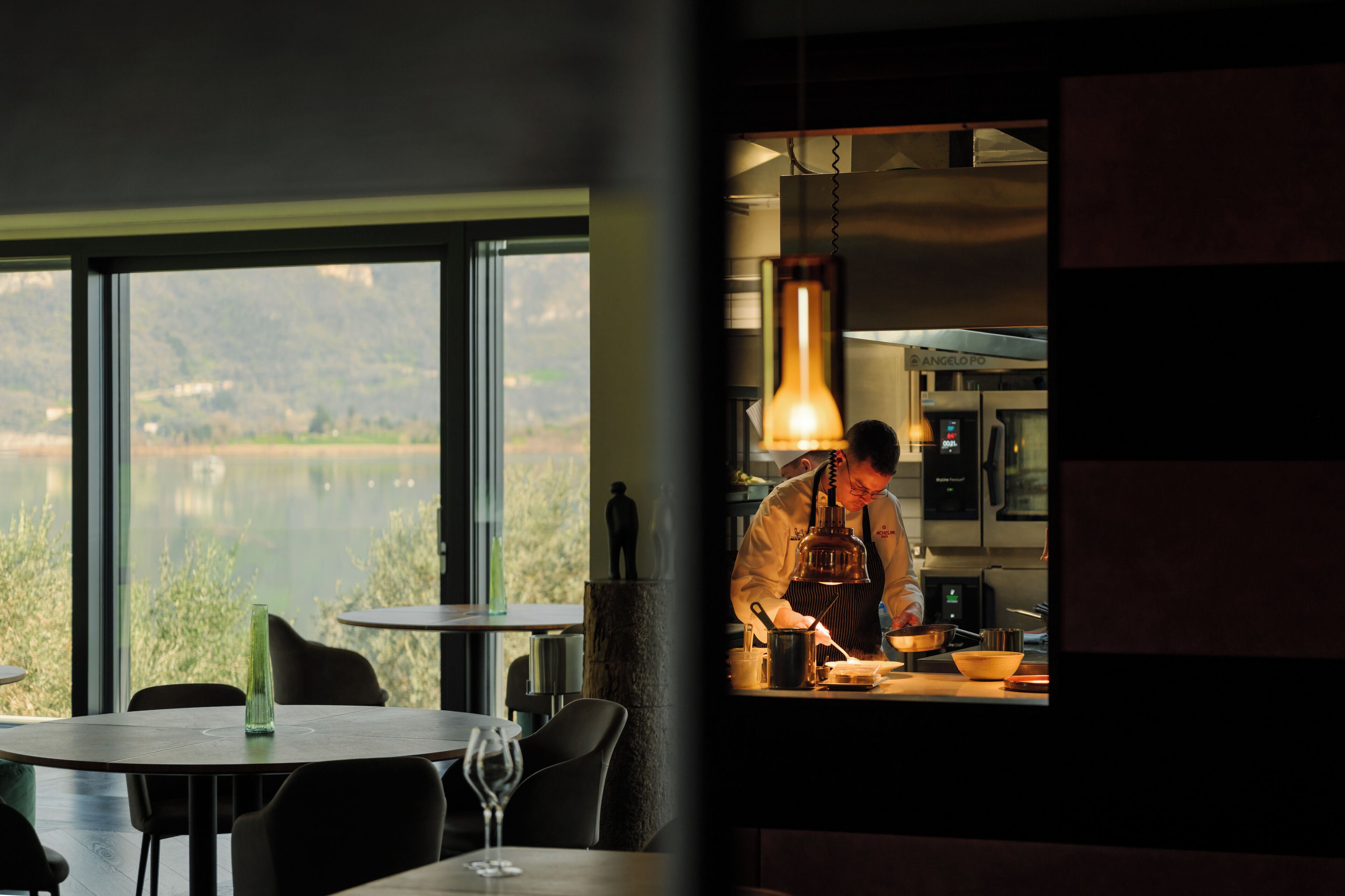 A restaurant with a view of a lake, a chef preparing food, and a dining area with tables and chairs.