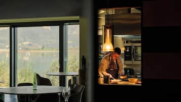 A restaurant with a view of a lake, a chef preparing food, and a dining area with tables and chairs.