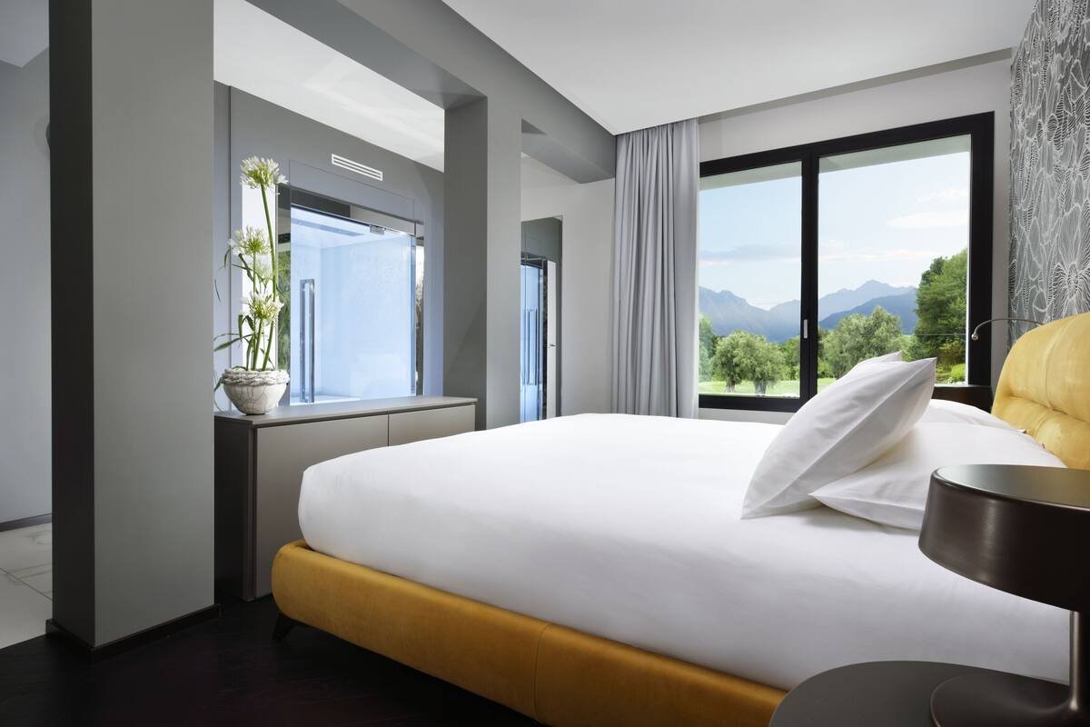 A modern hotel room with a large bed, a desk with a potted plant, and a view of mountains through large windows.