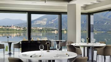 A modern dining area with round tables and chairs, large windows offering a view of a lake and mountains.