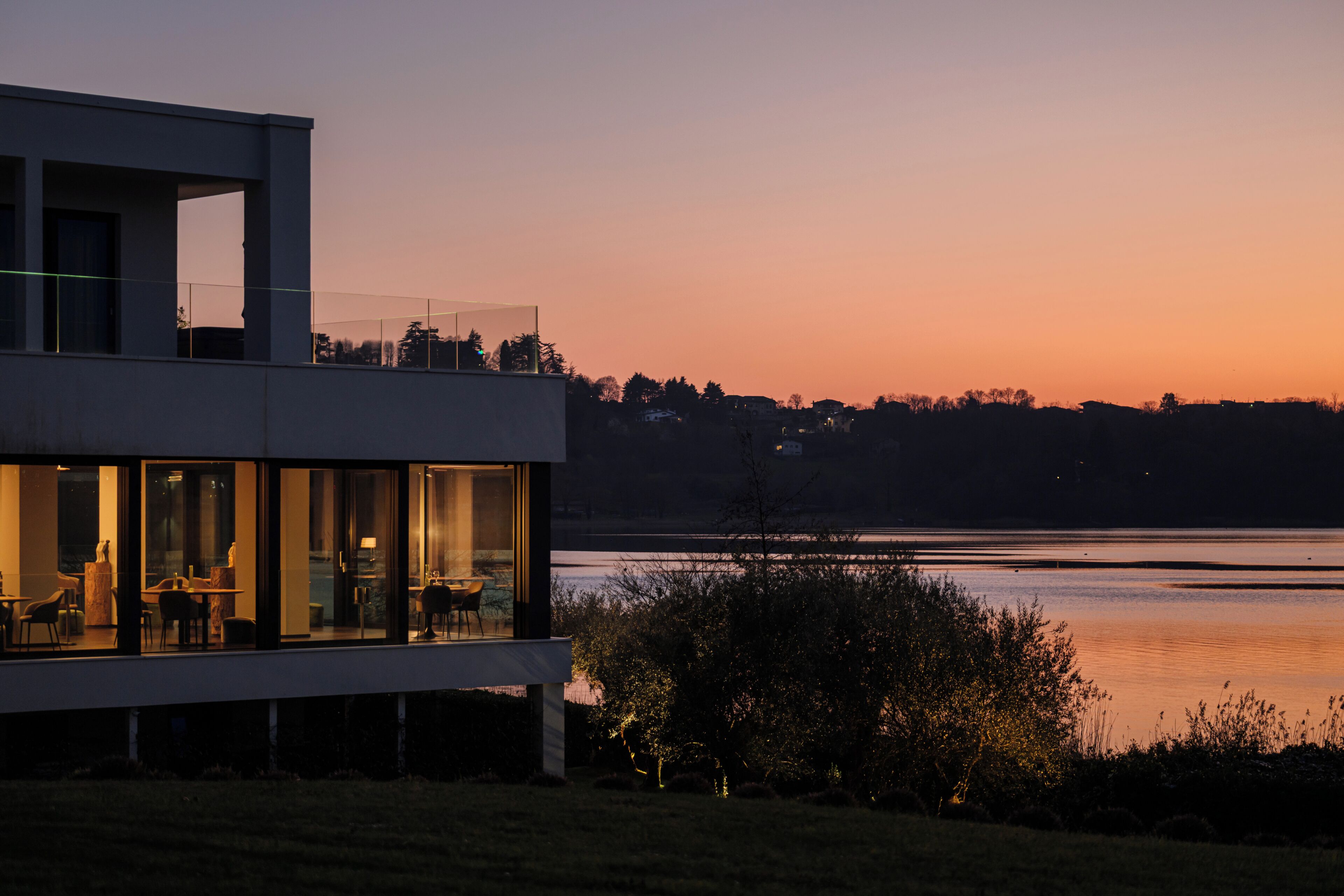 A modern building with large windows, a flat roof, and a view of a lake at dusk.