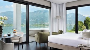 Hotelkamer met een groot bed, een eettafel met theeservies, twee stoelen en uitzicht op bergen en een meer.