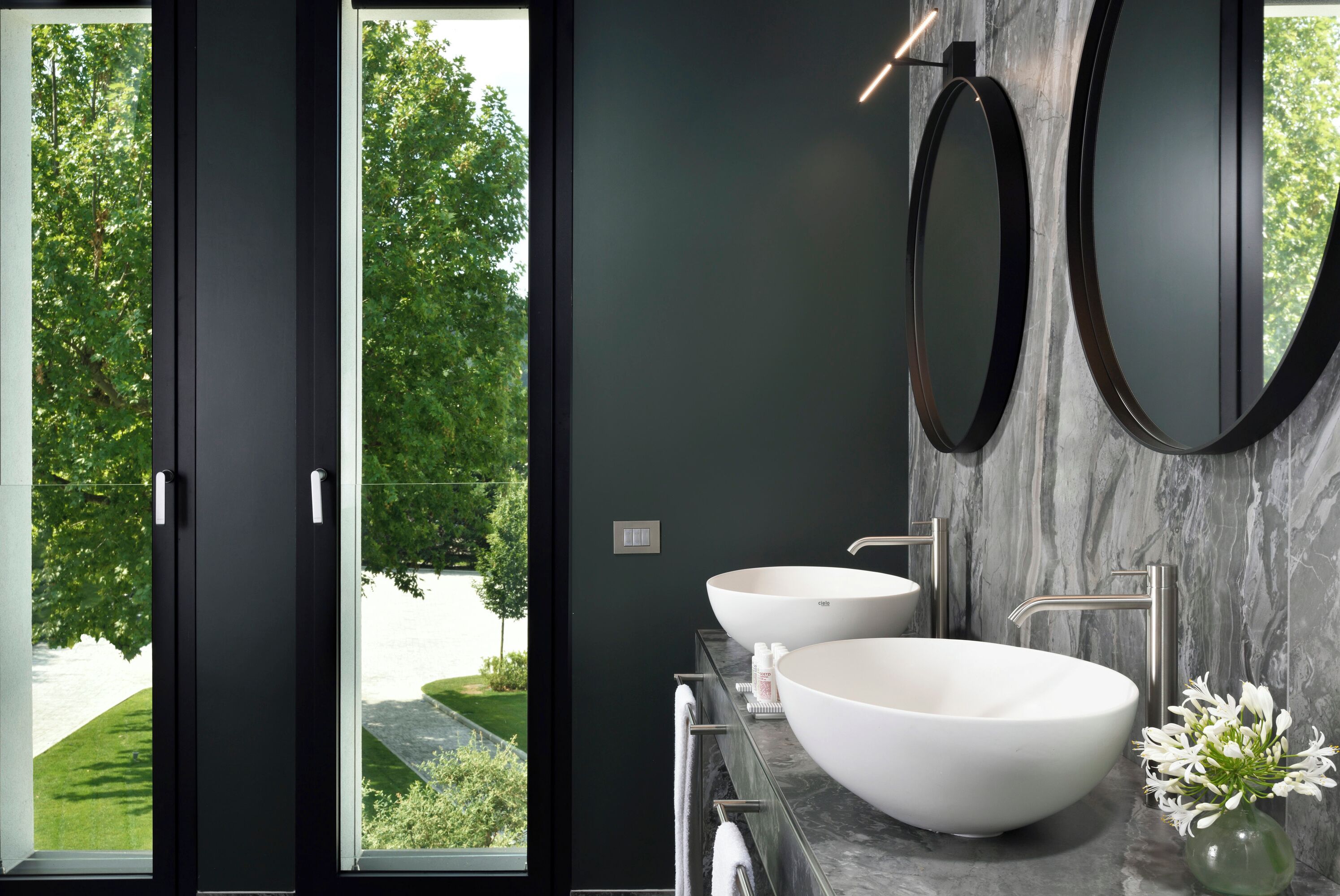 Un bagno moderno con due ampi lavandini bianchi, un piano di lavoro in marmo e vista sul verde attraverso una porta di vetro.
