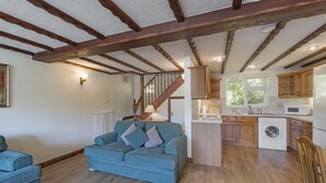 A spacious living area with a blue sofa and wooden flooring, leading to a kitchen with wooden cabinets and a washing machine. - A spacious 2 bedroom property (SALTASH)