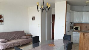 A living room with a glass dining table, black chairs, a sofa, and a kitchenette with a microwave and cabinets. - Duplex T3 for 6, beach nearby, private parking (Le Barcarès)