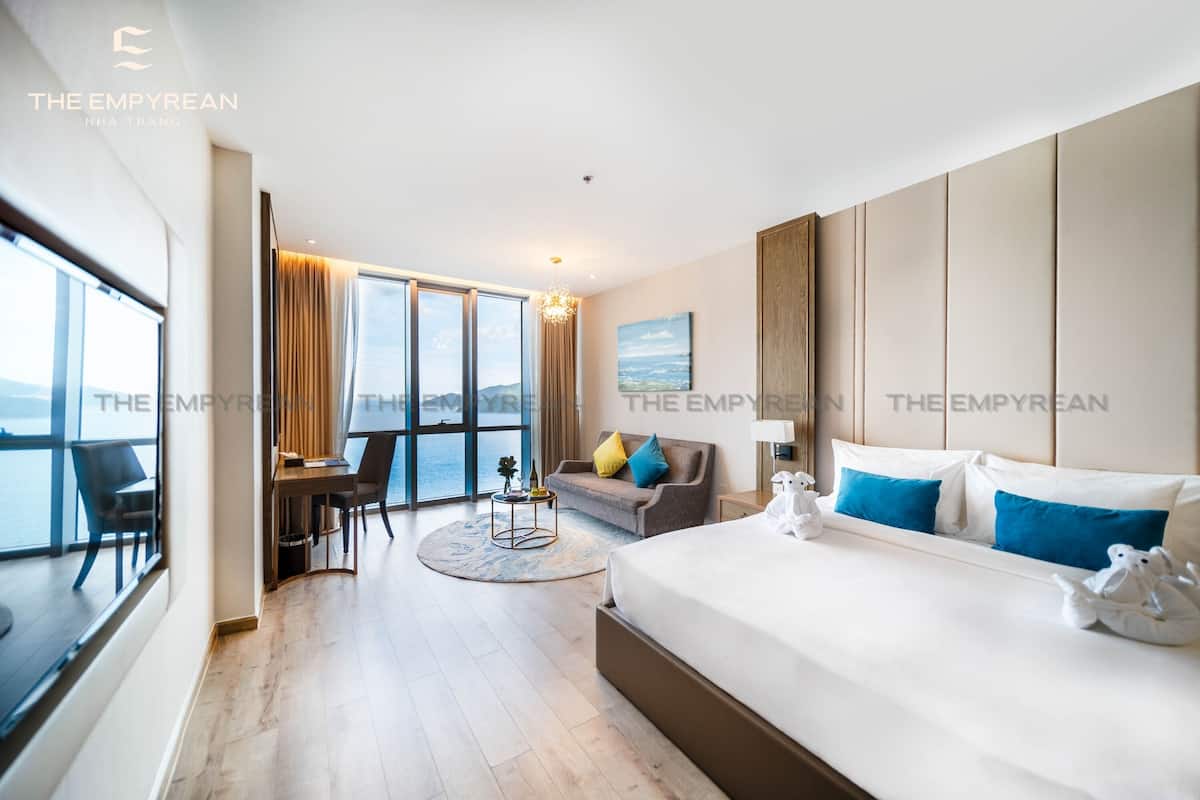 A modern hotel room with a large bed, a desk, and a sofa. There is a view of the sea through large windows.