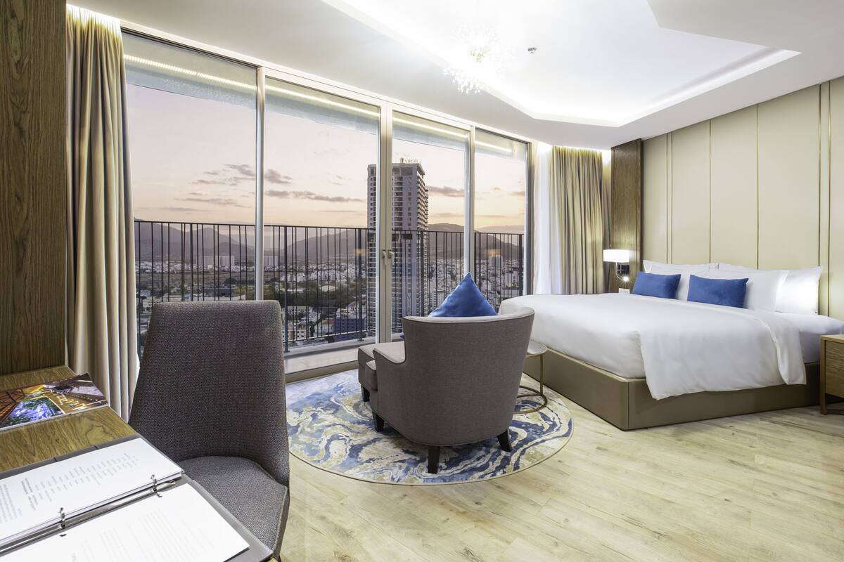 A hotel room with a large window, a bed, a chair, a desk, and a view of a cityscape.