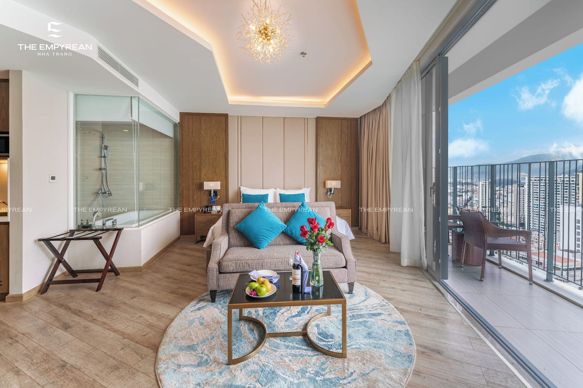 A modern hotel room with a balcony, a shower, a sofa with blue cushions, and a coffee table with fruit and a vase.