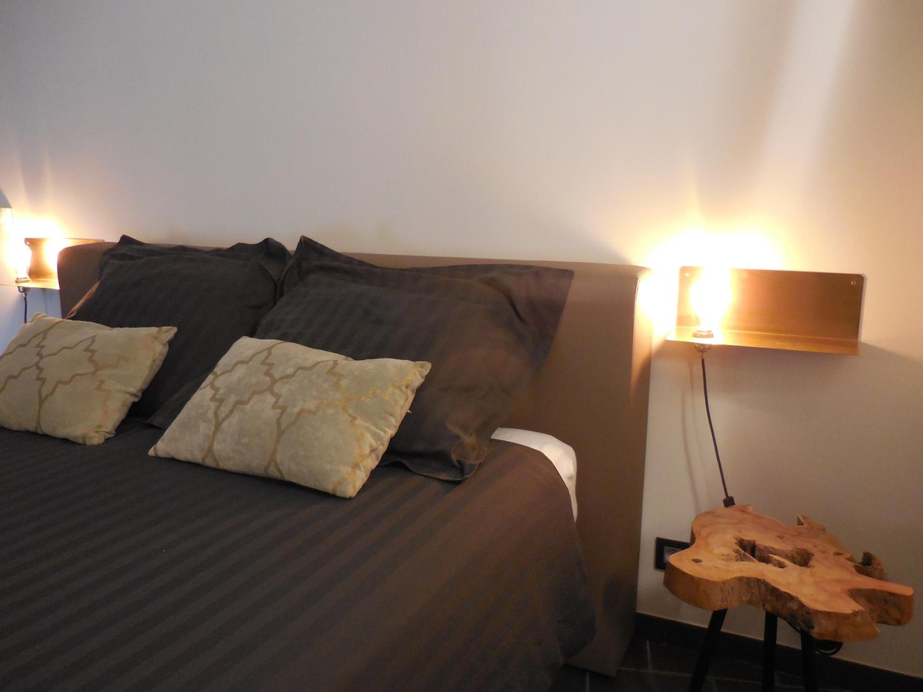 Un lit avec une literie sombre et des oreillers à motifs, une lampe fixée au mur et une guitare en bois posée sur un support.