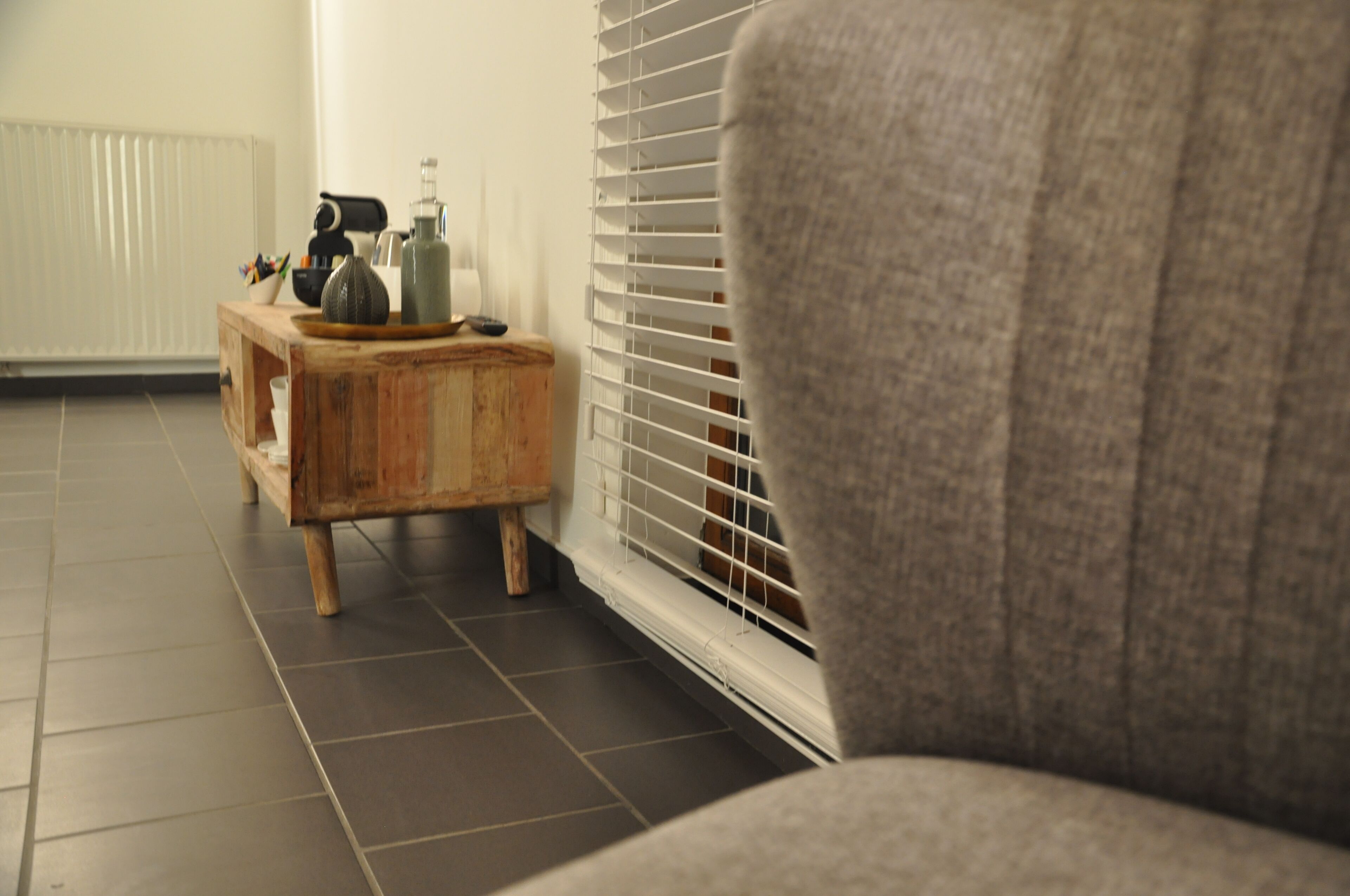 Una habitación con un sillón gris, una mesita de madera con un florero y un control remoto, y un radiador.