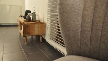 Una habitación con un sillón gris, una mesita de madera con un florero y un control remoto, y un radiador.