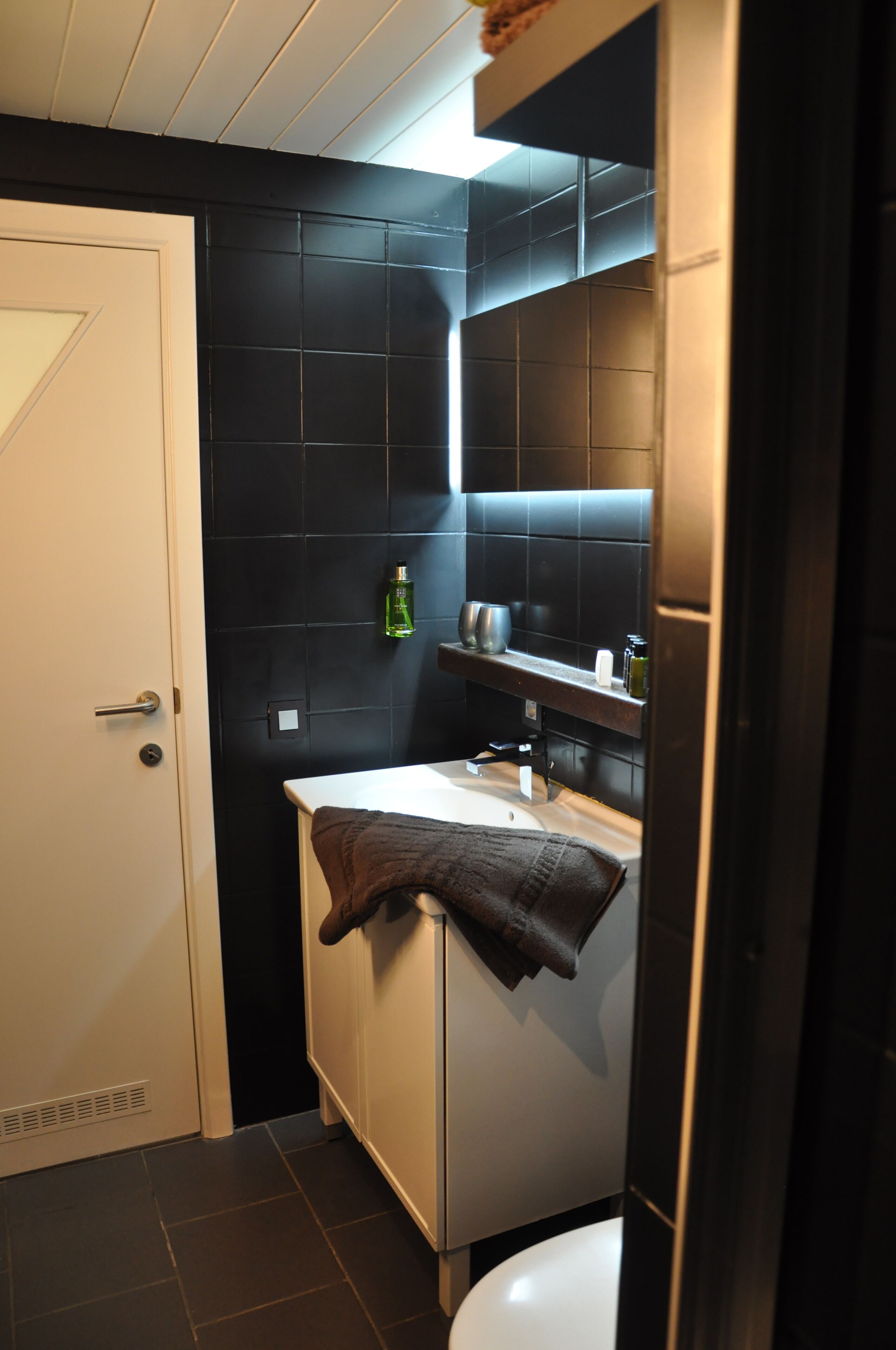 A compact bathroom with a white toilet, a small sink, and a mirror above it. There is a towel hanging on the wall and a bottle of hand soap on the shelf.