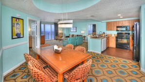 A modern kitchen with a black refrigerator, microwave, and oven. There is a dining area with a wooden table and patterned chairs. The walls are painted teal, and there is a large window in the background.