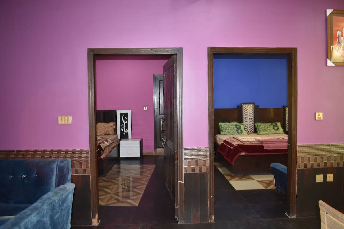 Une chambre d’hôtel avec un mur violet, un lit avec une literie verte et rouge, un fauteuil bleu et une armoire en bois.