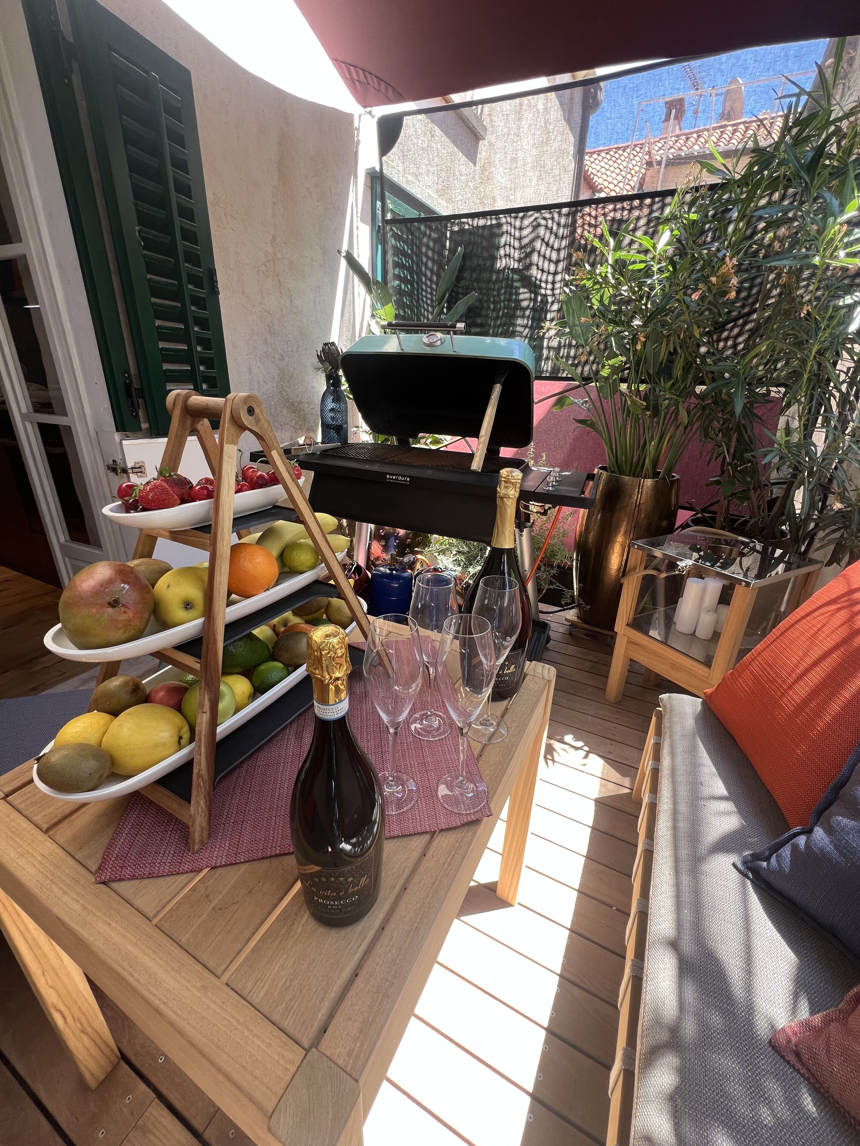 A wooden deck with a grill, fruit platter, wine glasses, and a bottle of Prosecco.