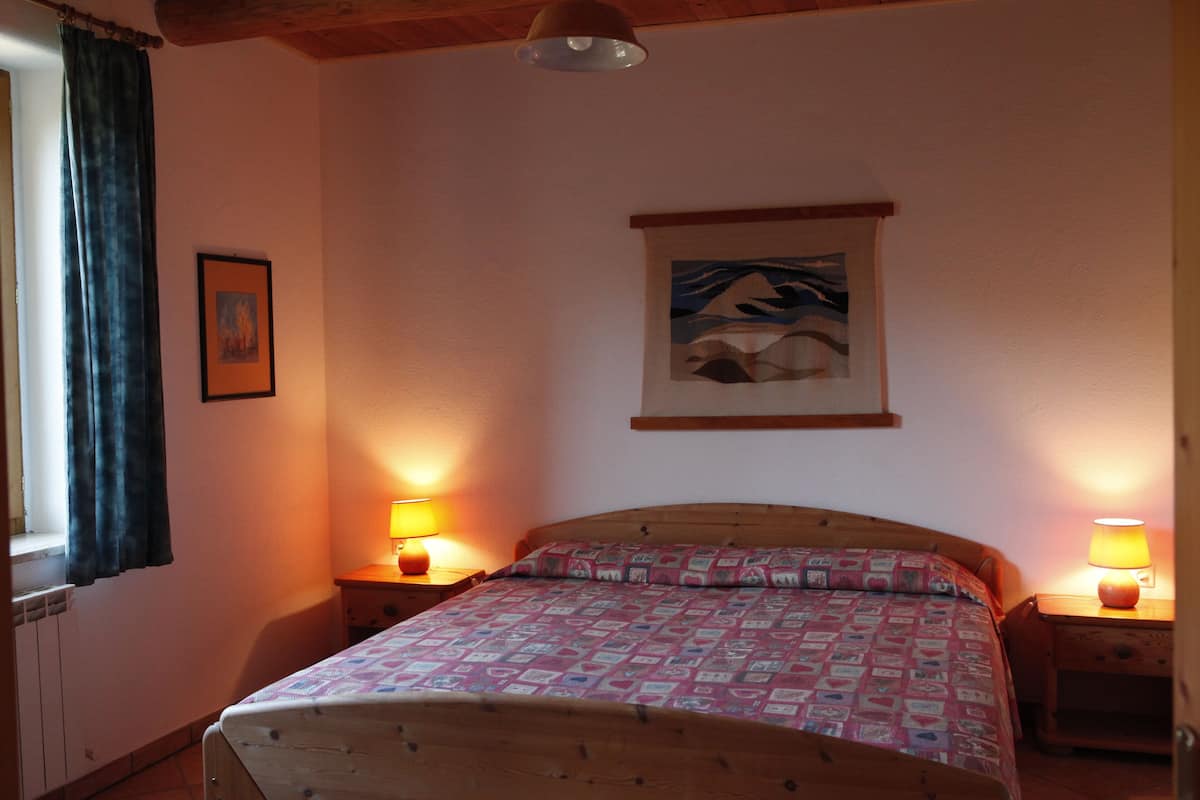 A bedroom with a wooden bed, two bedside lamps, a framed picture on the wall, and a window with curtains.