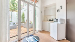 Cocina moderna con armarios blancos, microondas y un refrigerador pequeño. Hay una puerta corrediza de vidrio que da a un patio exterior con un sofá gris y un jardín pequeño.