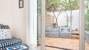 Una sala moderna con un sofá y una mesita, una puerta corrediza de vidrio que da a un patio exterior con piso de ladrillo y plantas en macetas.