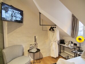 Ein modernes Hotelzimmer mit Fernseher, der ein Drama zeigt, einer Sitzecke mit einem Sessel, einem kleinen Tisch mit einem Tablett mit Gläsern, einem an einem Hängestuhl befestigten Bademantel und einem Nachttisch mit Lampe und Wasserkocher.