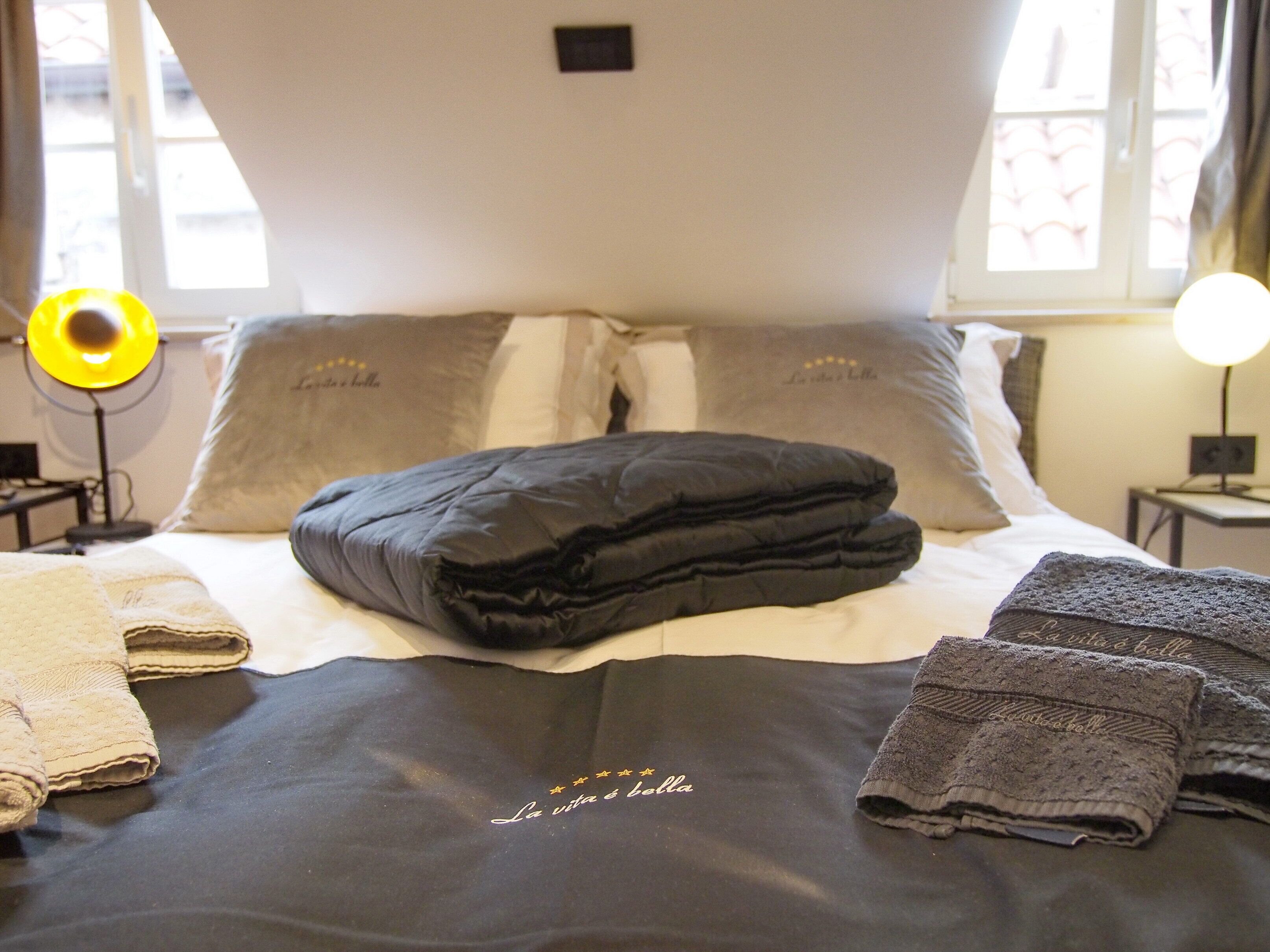 A neatly made bed with a black comforter and pillows, a bedside lamp, and a window with curtains.