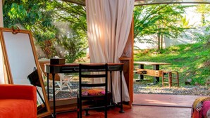 A room with a wooden desk, a chair, and a bed. There is a window with curtains, and a view of greenery outside. - Gadiamb City- Glamping (Saint-Pierre)