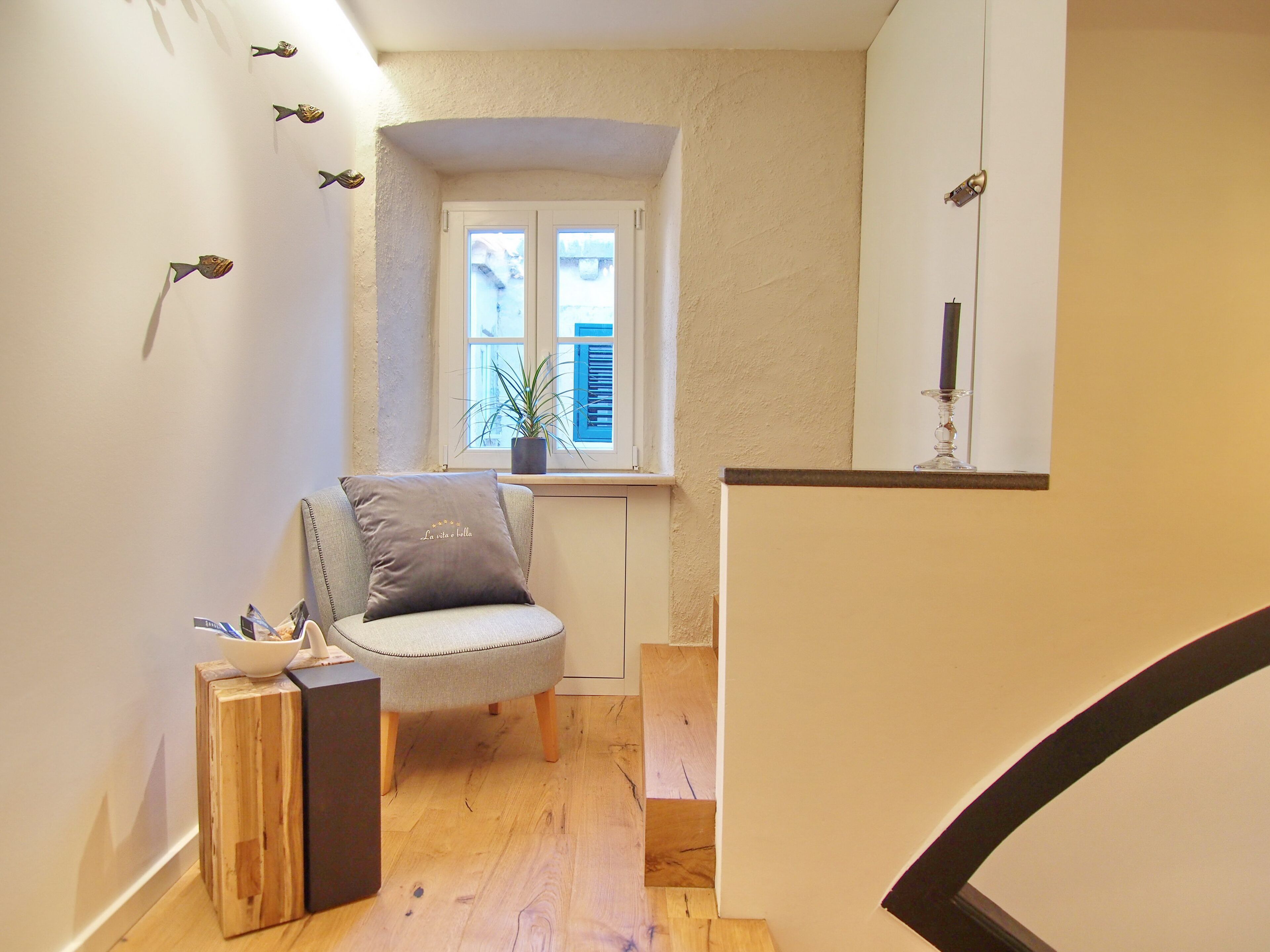 A cozy room with a wooden floor, a grey upholstered chair with a cushion, a small table with a bowl and a candle, and a window with a plant.
