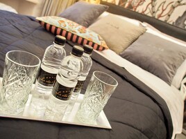 Uma cama com edredom preto e várias almofadas, um tabuleiro com quatro copos e três garrafas de água, e uma almofada decorativa com padrão floral.