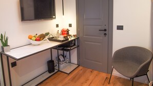 Una habitación pequeña y moderna con piso de madera, un escritorio blanco con un frutero, una cafetera roja y una silla gris.