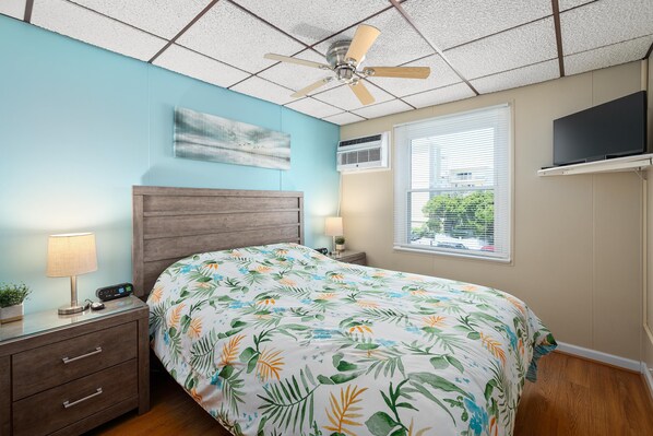 A bedroom with a bed, nightstand, lamp, ceiling fan, air conditioning unit, window, and a mounted TV.