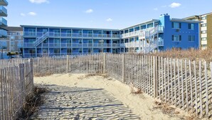 A beachfront hotel with a blue facade, multiple balconies, and a sandy pathway leading to the entrance. - Oceanfront condos with 3 night minimum rentals and linens included! (Ocean City)