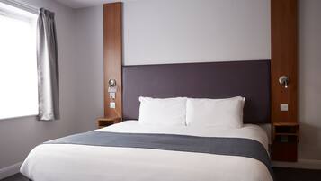 Una cama bien hecha con una manta gris, una mesita de noche de madera con una lámpara y un enchufe en la pared.