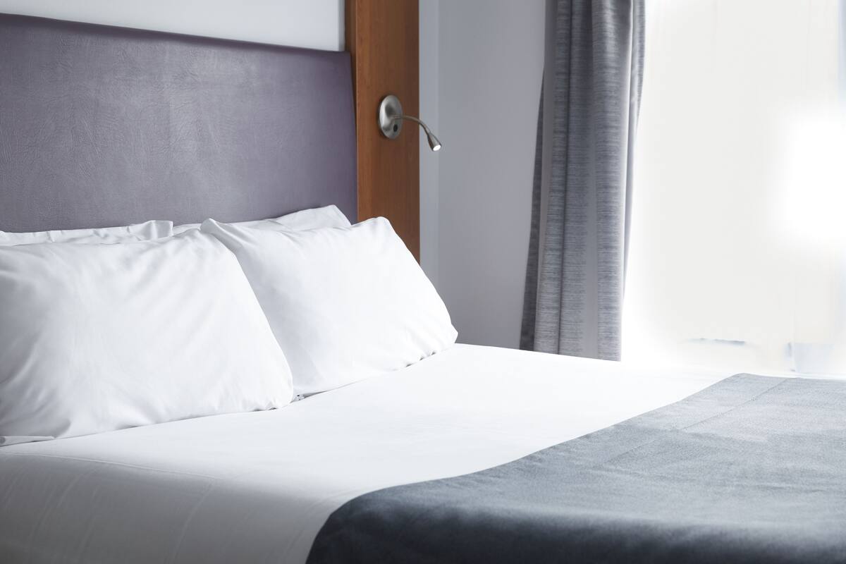 A neatly made bed with white linens and a grey throw blanket, a wooden headboard, and a window with grey curtains.
