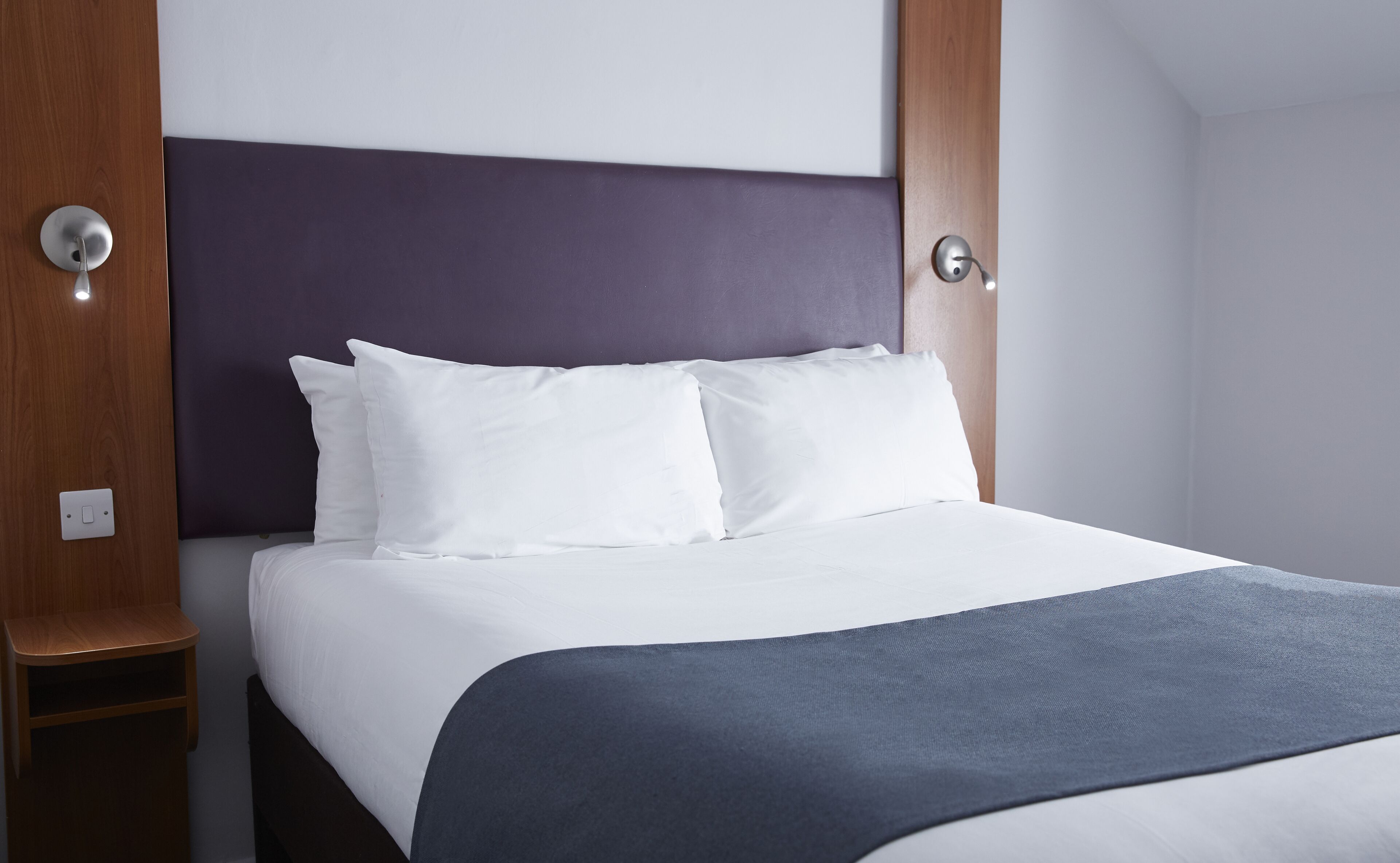 A neatly made bed with white linens and a dark blue throw blanket, a wooden nightstand, and a modern headboard.