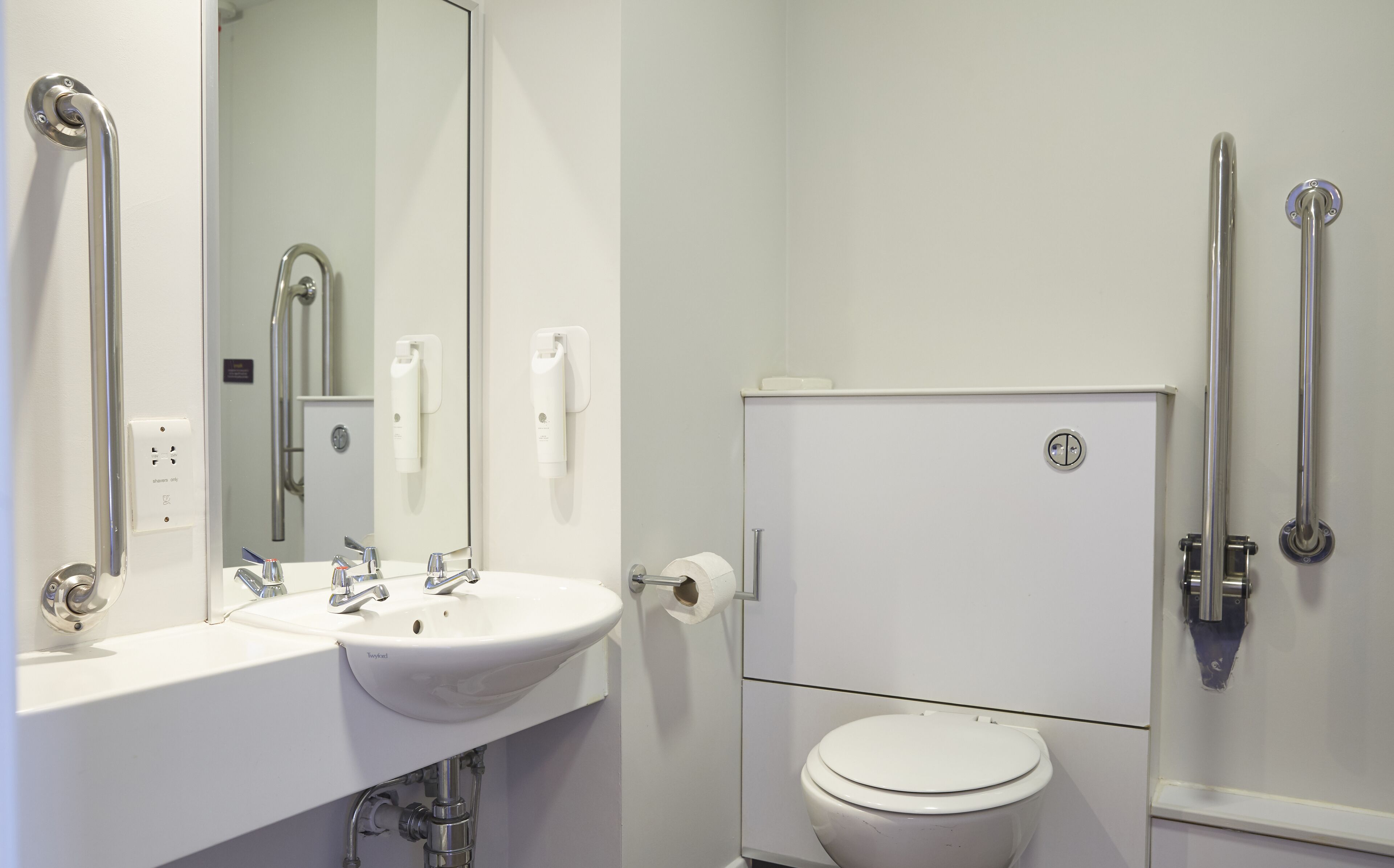 Un baño moderno con lavamanos, espejo y retrete blancos. Hay barras de apoyo en las paredes.