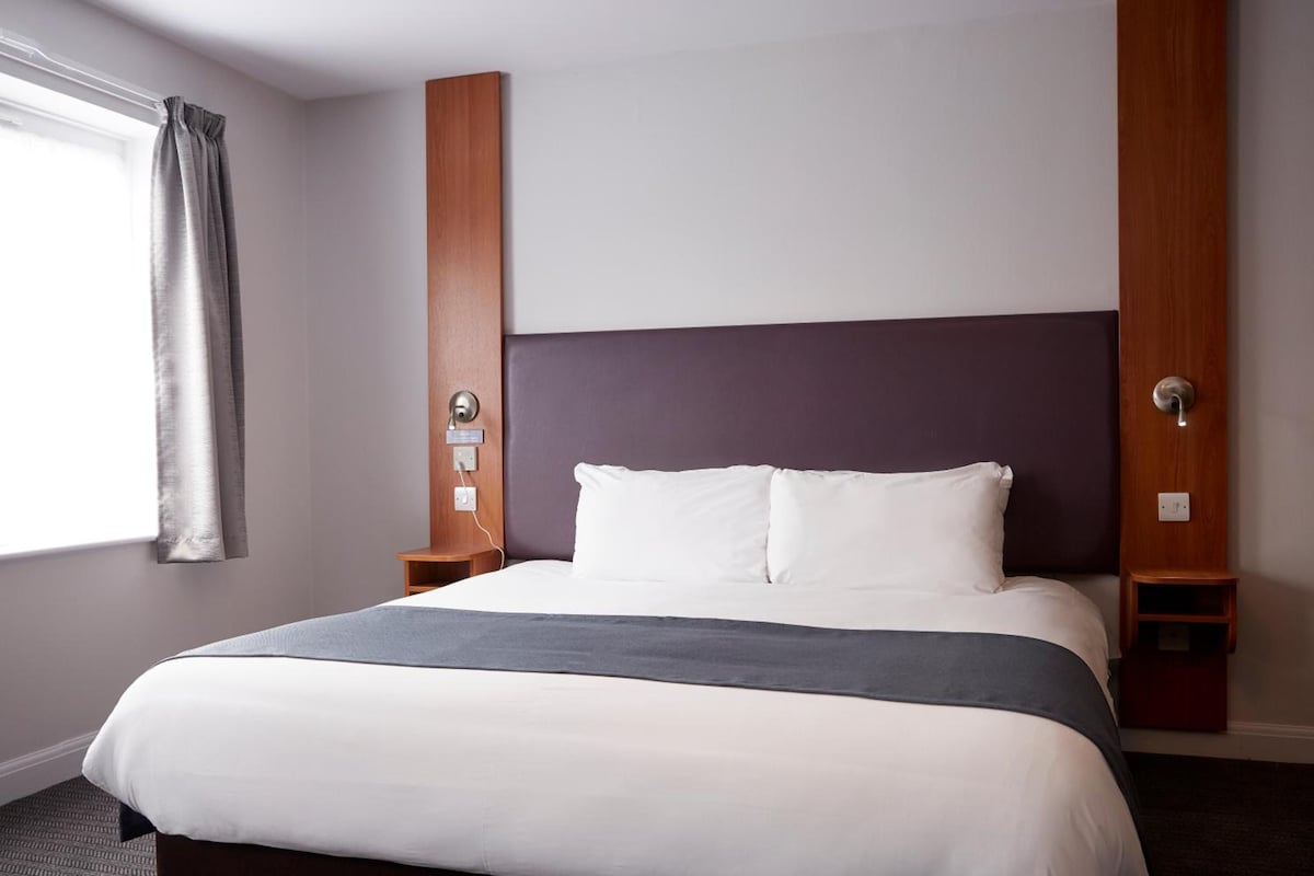 A hotel room with a large bed, a wooden headboard, a nightstand, and a window with curtains.