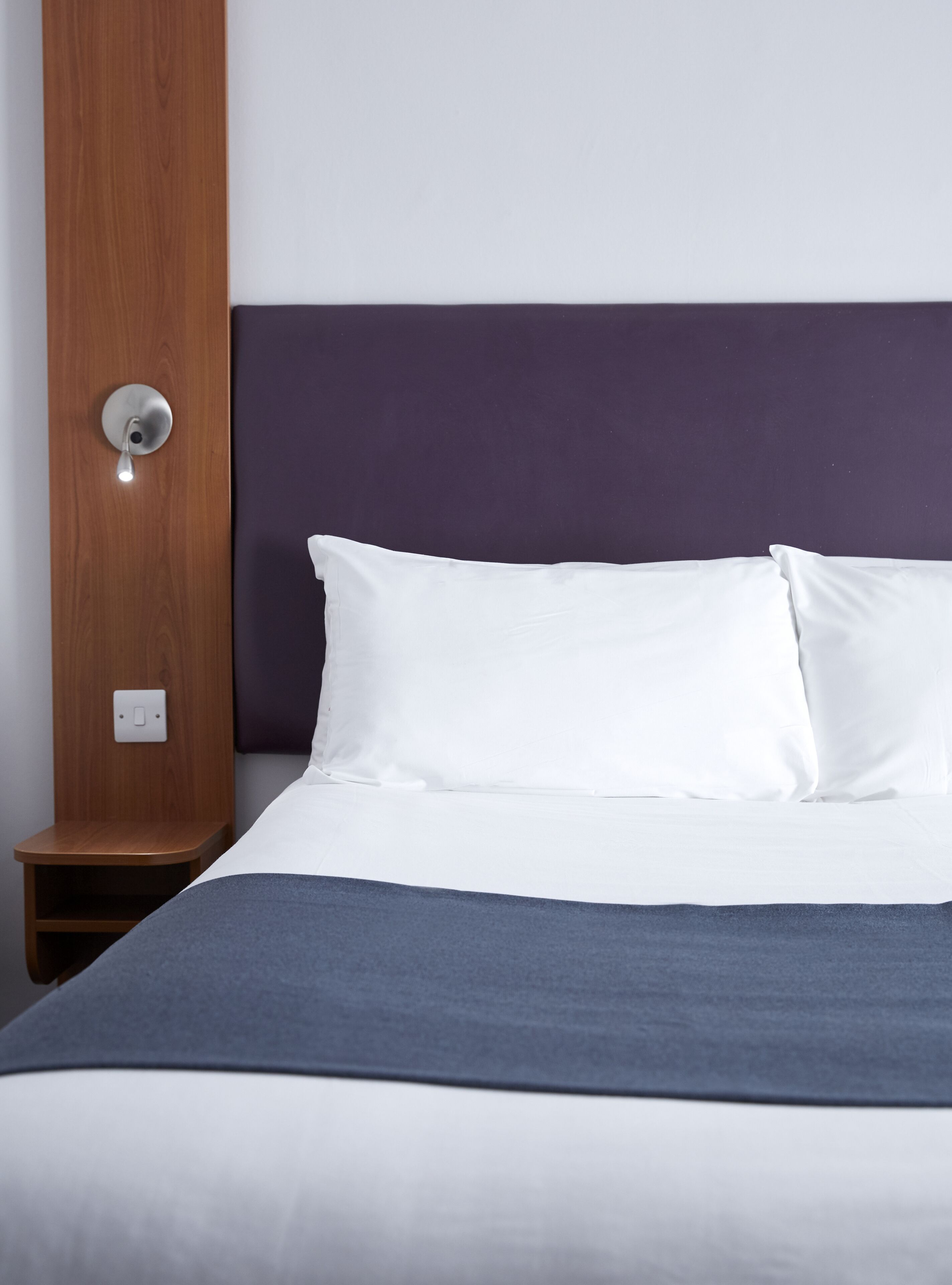 A neatly made bed with white linens and a blue throw blanket, a wooden nightstand with a light switch, and a purple headboard.