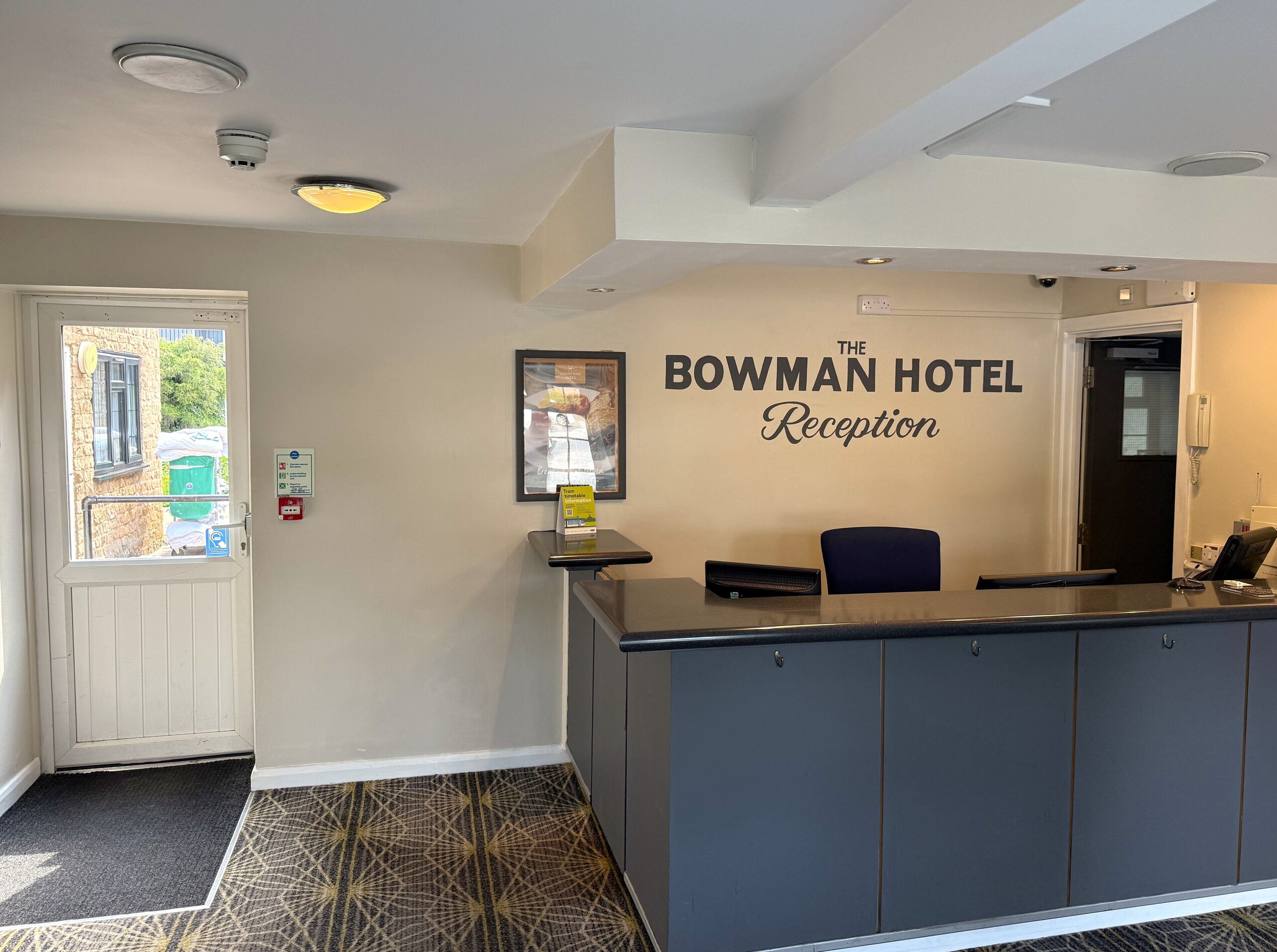 A hotel reception area with a reception desk, a framed welcome sign, a door, and a window with a view of the outside.