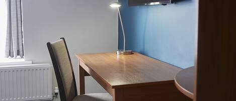 A room with a wooden desk, a chair, a desk lamp, a television mounted on the wall, and a window with curtains.