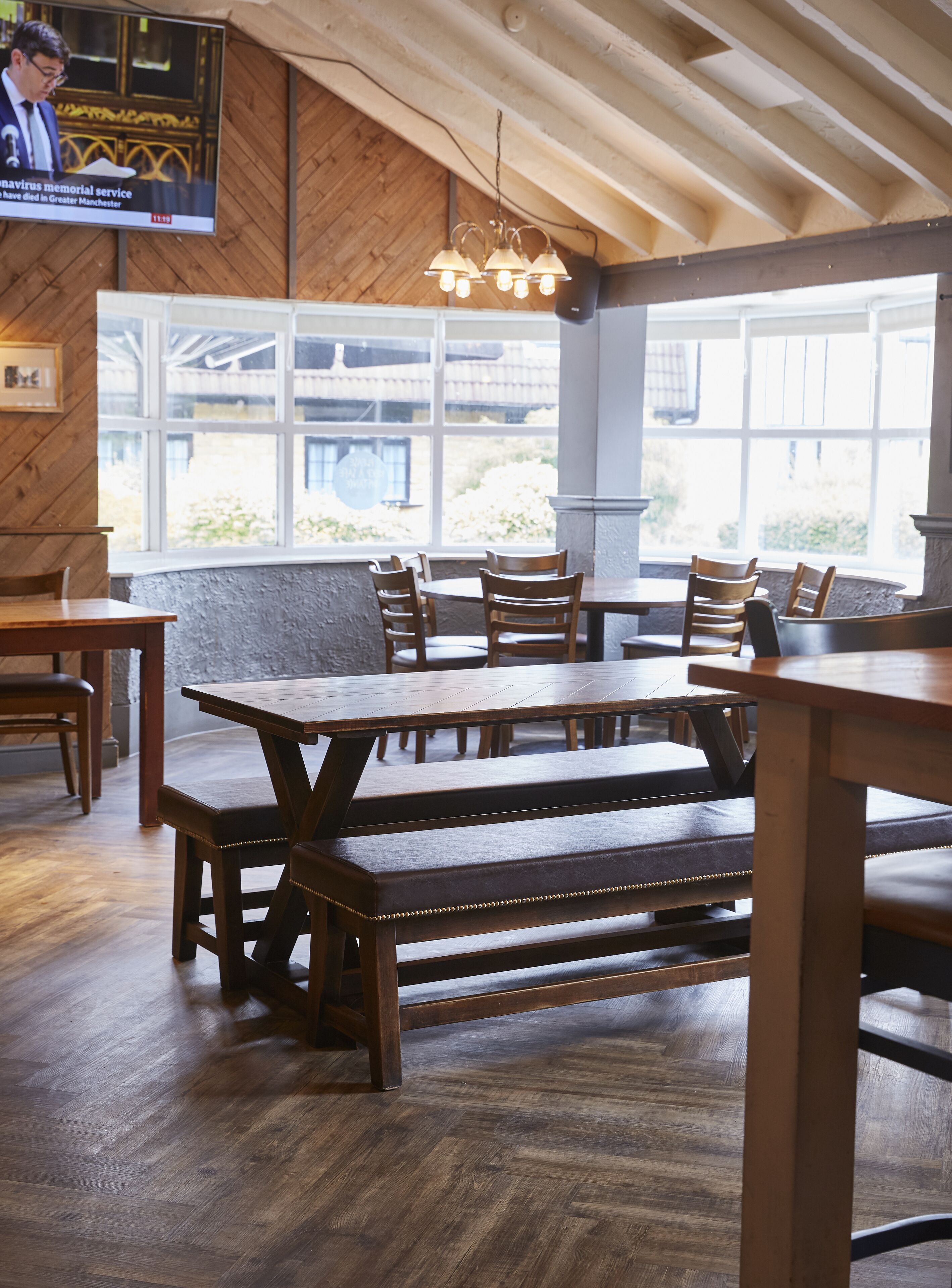 A restaurant interior with wooden tables and benches, large windows, and a television screen displaying news.