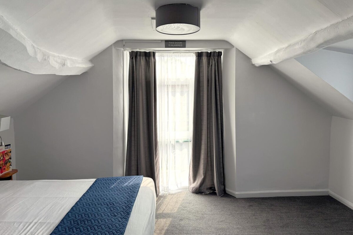 A hotel room with a bed, a window with curtains, a ceiling light, and a sign above the window.