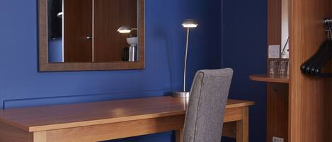 A room with a wooden desk, a chair, a mirror, a floor lamp, and a small table with glasses.