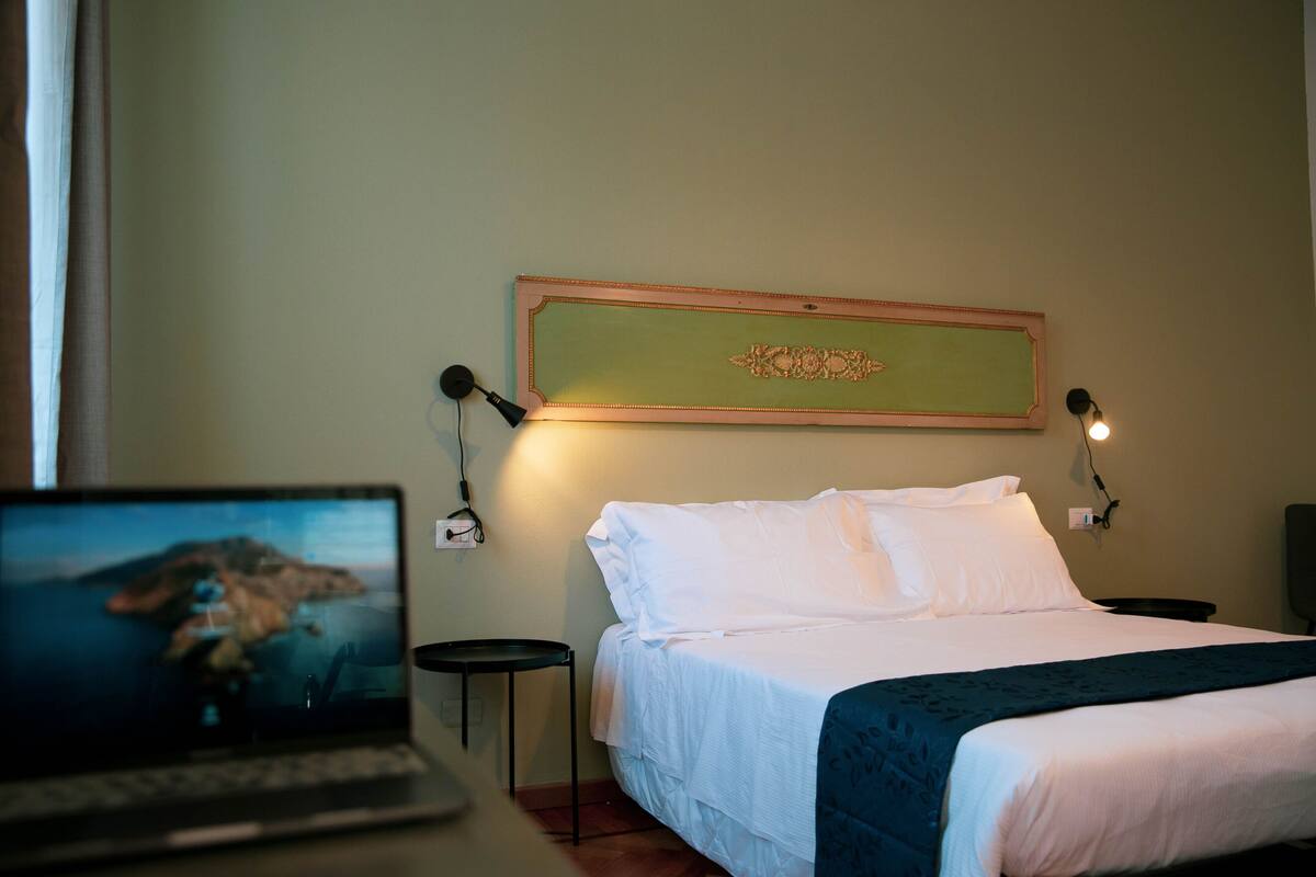 Un lit bien fait, avec du linge de lit blanc, un ordinateur portable posé sur une table de chevet et un tableau encadré accroché au mur.