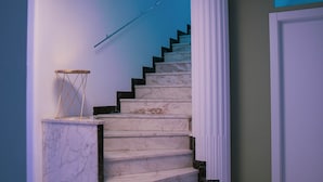 A marble staircase with a white door at the top, a small table with a geometric design, and a blue-lit ceiling. - Palazzo Pasta Malpensa (Gallarate)