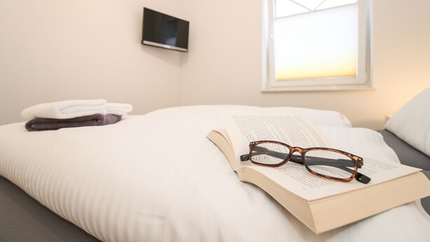 Ein ordentlich bezogenes Bett mit einem Buch und einer Brille darauf, ein Fenster mit Tageslicht und ein Fernseher an der Wand.